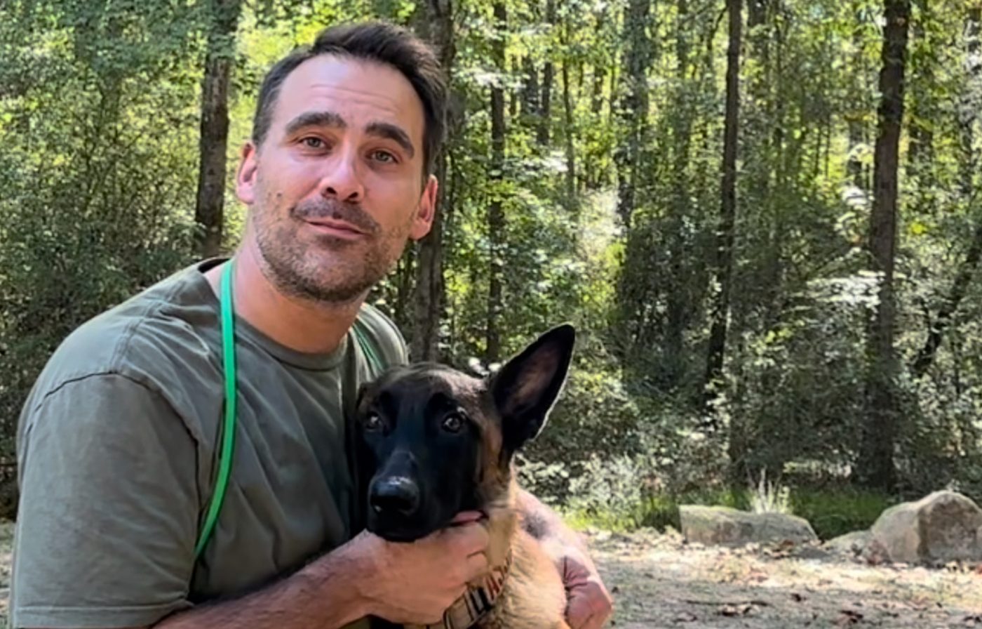 Joel Harrison crouched in the woods with a young shepherd-type dog leaning calmly into him.