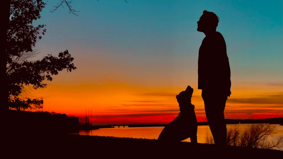 Silhouette of a person and a seated dog looking at each other against a vivid sunset over water.